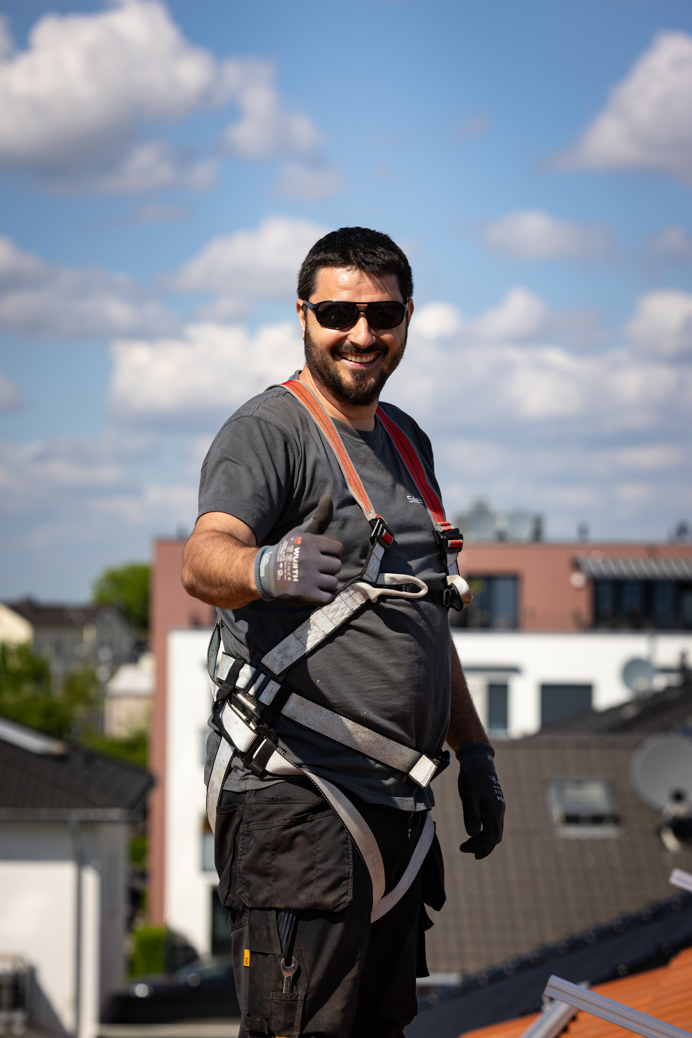 Ein Mann mit Sonnenbrille und Sicherheitsgeschirr lächelt und zeigt einen Daumen hoch auf einem Dach.