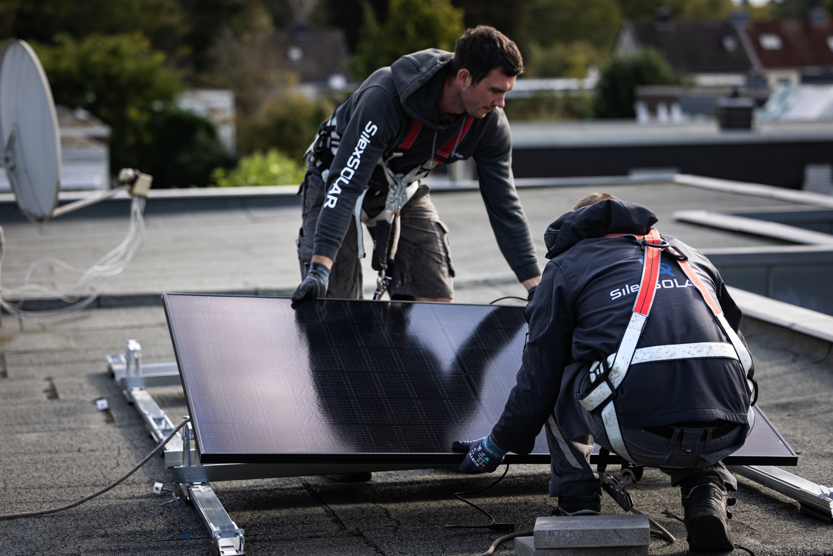 Zwei Arbeiter in Sicherheitsgurten installieren ein schwarzes Solarmodul auf einem flachen Dach.