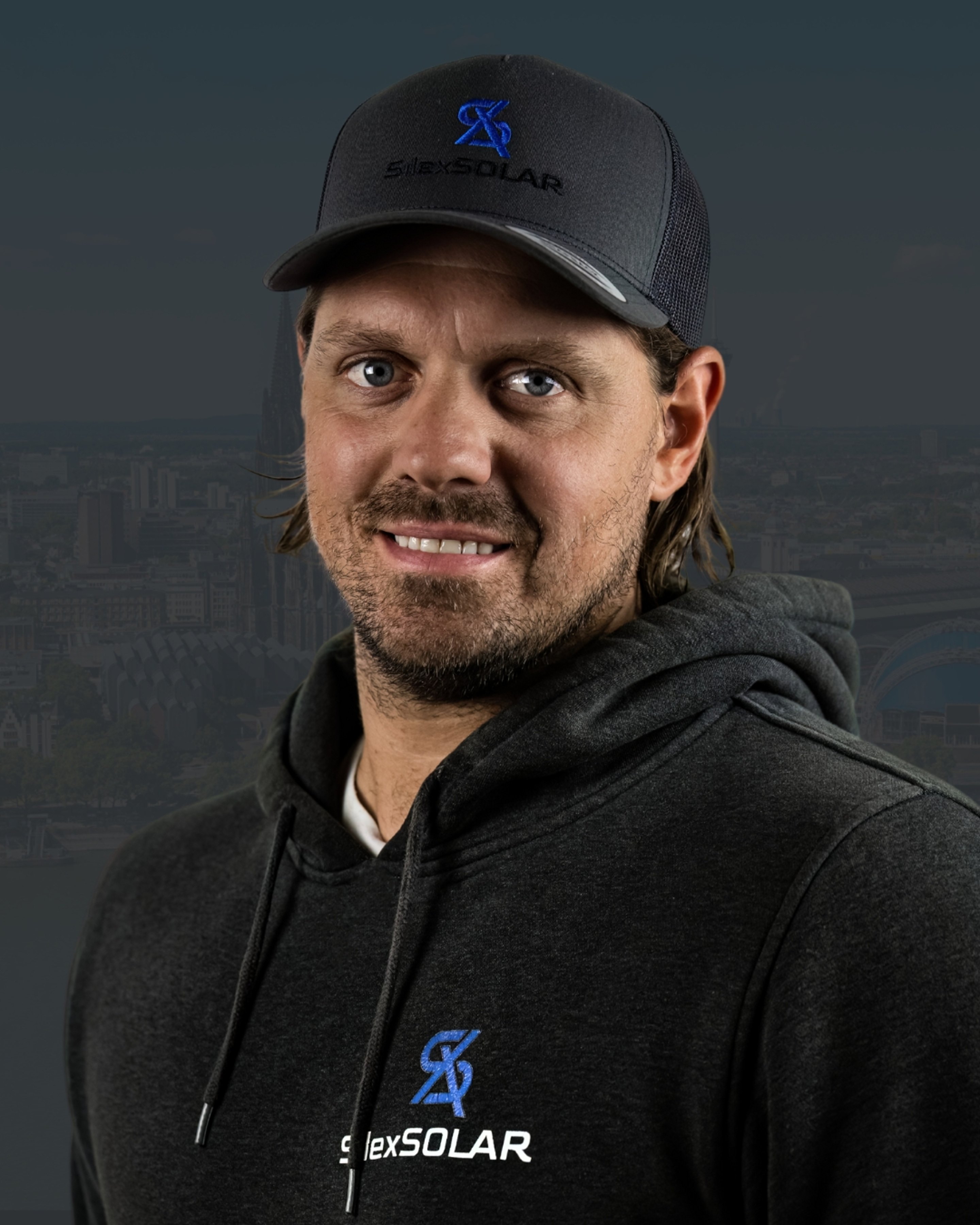 Mann mit Bart, dunkler Mütze und Kapuzenpullover mit „Slex SOLAR“-Logo lächelt.