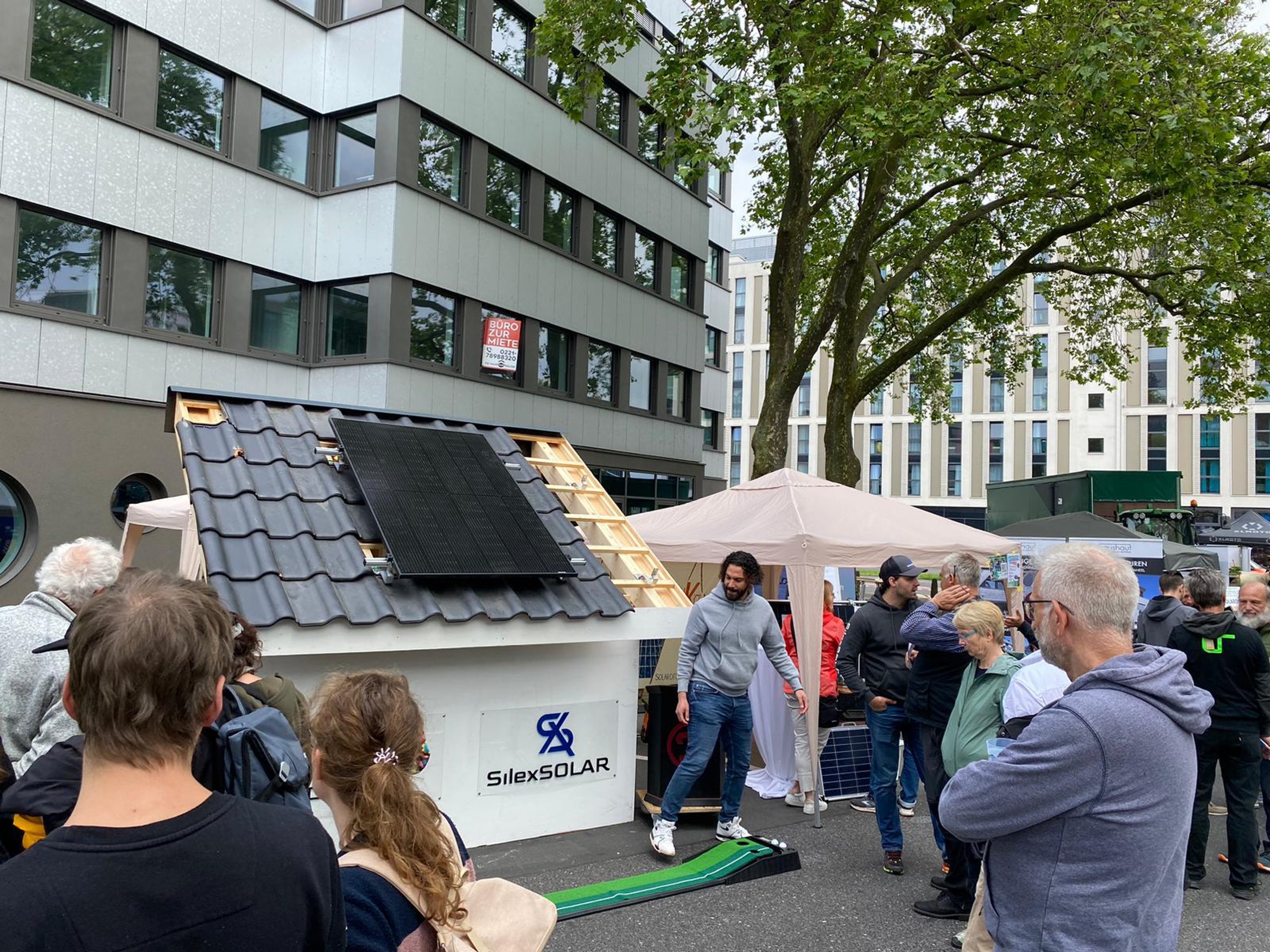 Ein Mann steht vor einem Modellhaus mit Solarmodulen auf dem Dach, umgeben von Menschen und einem Zelt.